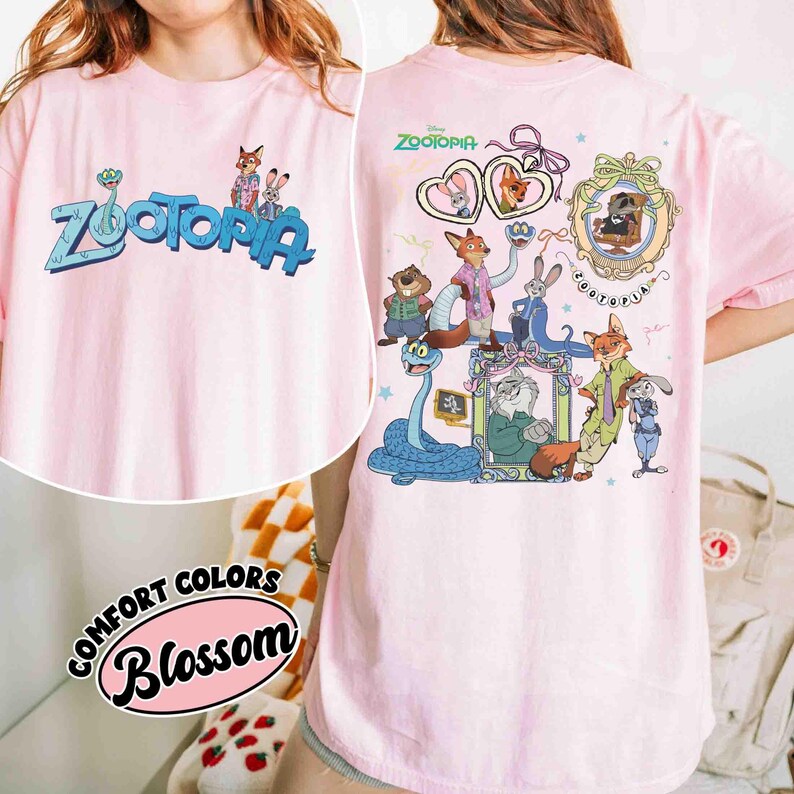 Puede incluir: Camiseta rosa claro con la palabra "Zootopia" en azul y personajes de dibujos animados. La parte trasera de la camiseta tiene gr&aacute;ficos adicionales de personajes de Zootopia. La camiseta est&aacute; etiquetada como "Comfort Colors Blossom".