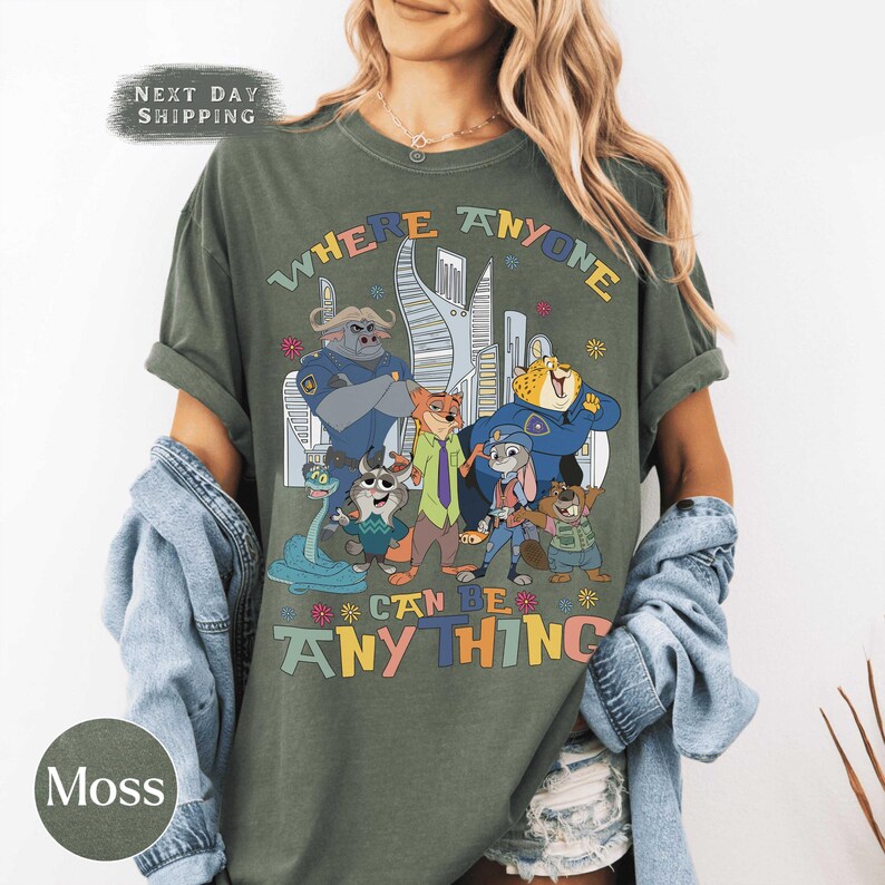 Puede incluir: Camiseta verde musgo con un gr&aacute;fico colorido de personajes de dibujos animados y el texto "WHERE ANYONE CAN BE ANYTHING". El dise&ntilde;o incluye un paisaje urbano y varias figuras de animales. La camiseta tiene mangas enrolladas.