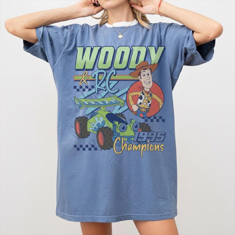 May include: Blue t-shirt featuring a graphic of Woody and RC from Toy Story. The design includes the text "WOODY & RC" and "1995 Champions". The shirt has short sleeves and a crew neck.