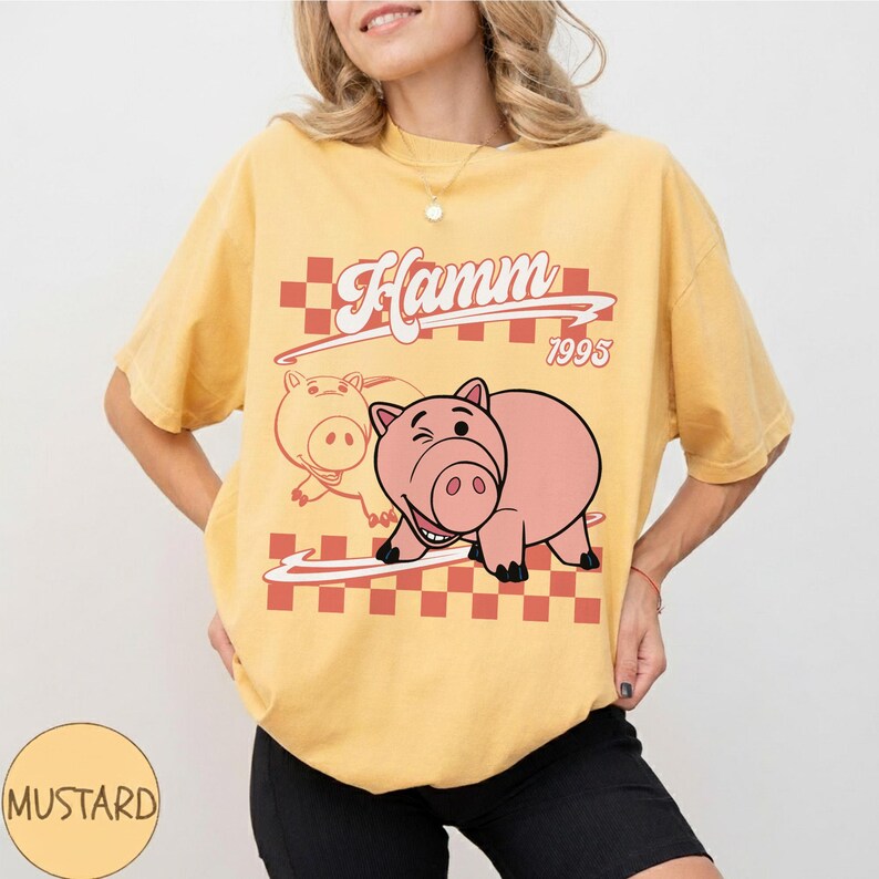 May include: Mustard-colored t-shirt featuring a graphic of Hamm from Toy Story. The design includes the word "Hamm" in a retro font, the year "1995", and a checkered pattern. The shirt is a relaxed fit.