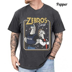 May include: Dark grey t-shirt with a cartoon graphic of two zebras. One zebra is dressed as a police officer, and the other is wearing a jacket. The text "ZEBROS + Zebras" is displayed in gold.