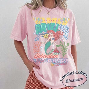 May include: A light pink short-sleeve t-shirt featuring a graphic print of Ariel and Flounder with the text Atlantica Ocean Tour 1991, Sebastian, and Hot Crustacean Band. The design includes coral and seaweed illustrations in blue, green, and yellow.
