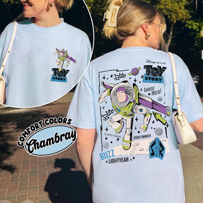 K&ouml;nnte beinhalten: Hellblaues T-Shirt mit einer Grafik von Buzz Lightyear aus Toy Story. Die Vorderseite hat ein kleines Bild, w&auml;hrend die R&uuml;ckseite ein gr&ouml;&szlig;eres Design mit dem Text "Galactic Hero!" und "Buzz + Lightyear" zeigt. Das Shirt ist mit "Comfort Colors Chambray" gekennzeichnet.