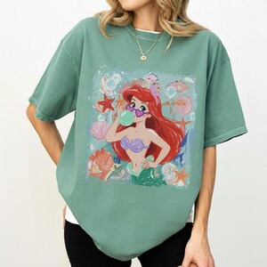 May include: A sage green short-sleeve t-shirt featuring a graphic print of a red-haired mermaid wearing heart-shaped sunglasses and blowing a bubble. The design includes various seashells, starfish, and bubbles surrounding the mermaid figure.