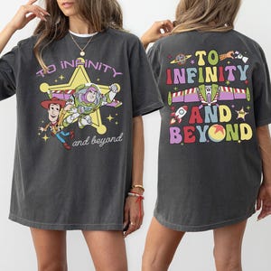 May include: Dark gray t-shirt featuring a graphic of Woody and Buzz Lightyear with the text "To Infinity and Beyond." The back of the shirt has the same phrase with a rocket, spaceship, and other colorful elements.