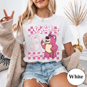 May include: White t-shirt featuring a pink graphic of Lotso, a bear character, with the text "Lotso 1995" in a pink checkered design. The bear is depicted with a cane and a friendly expression.