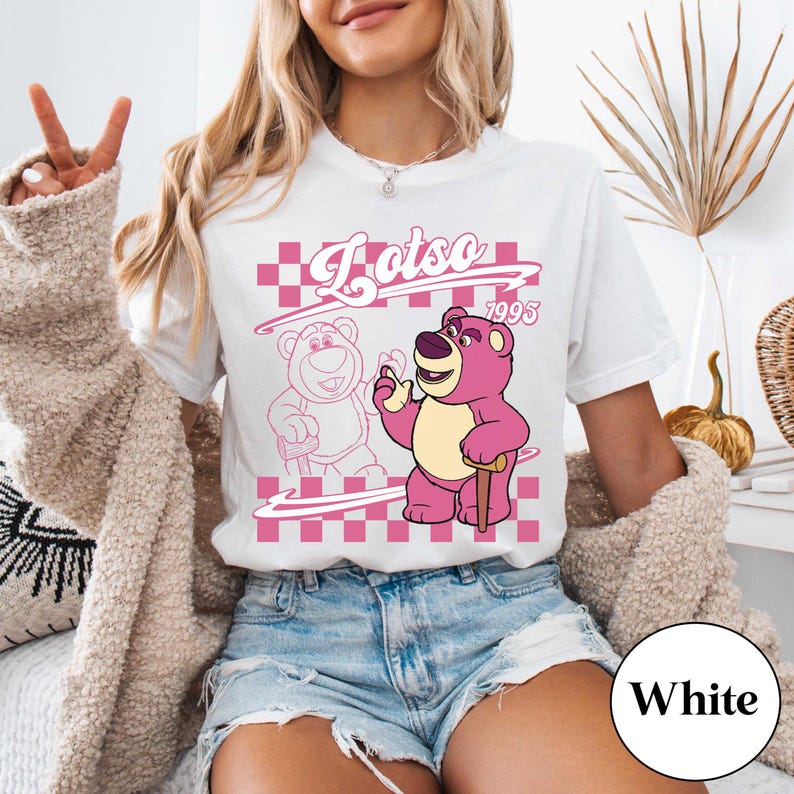 May include: White t-shirt featuring a pink graphic of Lotso, a bear character, with the text "Lotso 1995" in a pink checkered design. The bear is depicted with a cane and a friendly expression.