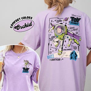 K&ouml;nnte beinhalten: Orchideenfarbenes T-Shirt mit einer Grafik von Buzz Lightyear aus Toy Story. Das Design enth&auml;lt den Text "Toy Story", "Galactic Hero!" und "Space Ranger". Das Shirt hat auch den Text "Comfort Colors" und "Orchid".