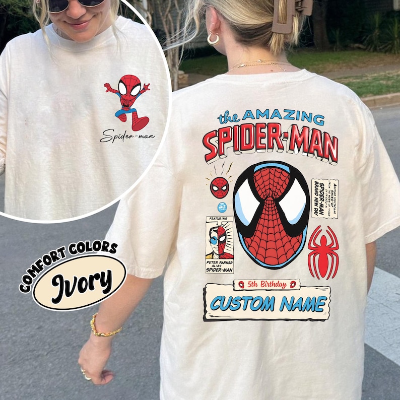 May include: Ivory-colored long-sleeve t-shirt featuring Spider-Man graphics. The front has a small Spider-Man cartoon and the text "Spider-man." The back displays "the AMAZING SPIDER-MAN" with Spider-Man's mask and other comic-style elements. Includes "CUSTOM NAME."