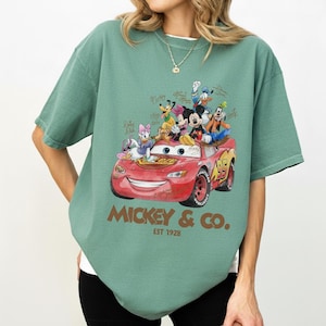 May include: Green t-shirt featuring a graphic of a red race car with Disney characters, including Mickey Mouse, Donald Duck, and Goofy. The text "Mickey & Co. Est 1928" is printed below the car.