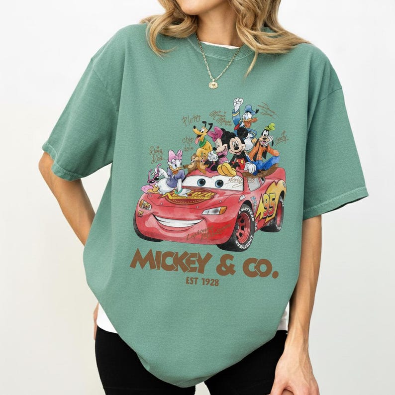 May include: Green t-shirt featuring a graphic of a red race car with Disney characters, including Mickey Mouse, Donald Duck, and Goofy. The text "Mickey & Co. Est 1928" is printed below the car.