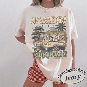 May include: Ivory-colored t-shirt with a safari-themed graphic. The design features a jeep with cartoon characters, the text "JAMBO!" and "KILIMANJARO SAFARI TOUR". The shirt is labeled "Comfort Colors".