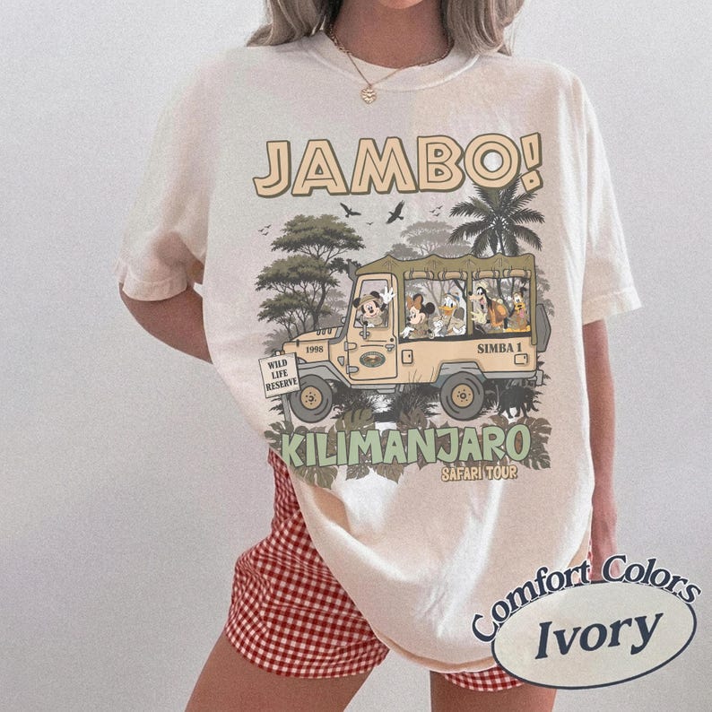 May include: Ivory-colored t-shirt with a safari-themed graphic. The design features a jeep with cartoon characters, the text "JAMBO!" and "KILIMANJARO SAFARI TOUR". The shirt is labeled "Comfort Colors".
