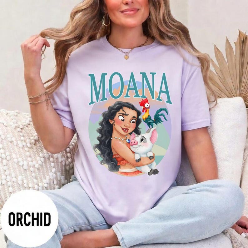 May include: A light purple t-shirt featuring the word "MOANA" in teal above a graphic of Moana, a pig, and a rooster. The shirt has a round neckline and short sleeves. The word "ORCHID" is printed on a white circle at the bottom left.