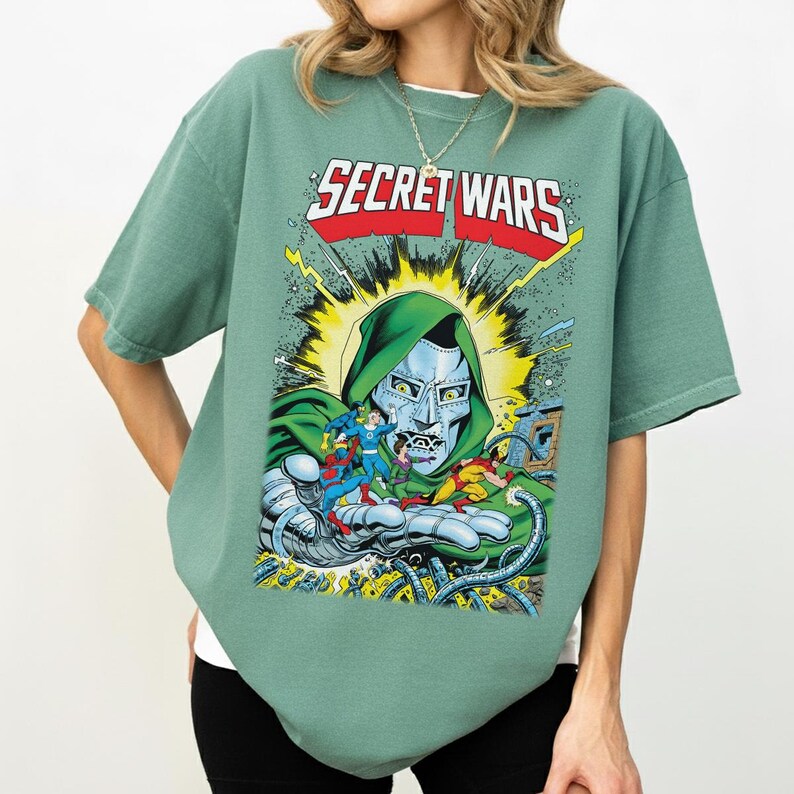 May include: Sage green t-shirt featuring a comic book graphic. The graphic includes the text "SECRET WARS" in red and white, along with a depiction of Doctor Doom and other comic book characters. The shirt has short sleeves and a crew neck.
