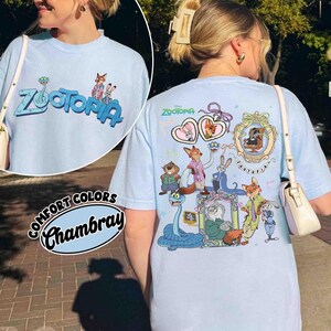 Puede incluir: Camiseta azul claro "Comfort Colors Chambray" con la palabra "Zootopia" y personajes de dibujos animados. La parte delantera muestra "Zootopia" en azul y verde. La parte trasera presenta varios personajes de dibujos animados.