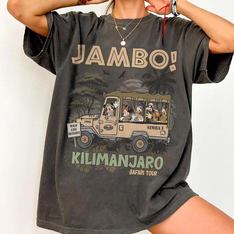 May include: Dark gray t-shirt with the word "JAMBO!" above a safari jeep illustration. The jeep features Mickey Mouse, Minnie Mouse, Donald Duck, and Goofy. The text "KILIMANJARO SAFARI TOUR" is below the jeep.