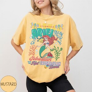 May include: A mustard yellow short-sleeve t-shirt featuring a graphic print of Ariel, Flounder, and Sebastian. The text reads Atlantica Ariel Ocean Tour 1991, Sebastian with the Hot Crustacean Band, with coral and seaweed illustrations in blue and red.