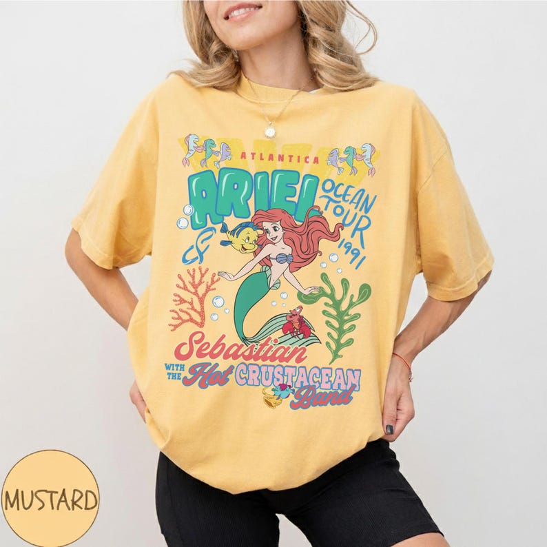 May include: A mustard yellow short-sleeve t-shirt featuring a graphic print of Ariel, Flounder, and Sebastian. The text reads Atlantica Ariel Ocean Tour 1991, Sebastian with the Hot Crustacean Band, with coral and seaweed illustrations in blue and red.