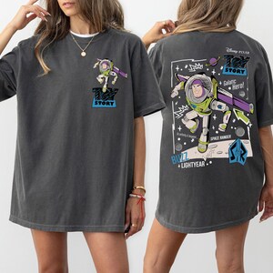 K&ouml;nnte beinhalten: Dunkelgraues T-Shirt mit einem Buzz Lightyear-Motiv aus Toy Story. Die Vorderseite zeigt ein kleines Buzz-Bild, w&auml;hrend die R&uuml;ckseite ein gr&ouml;&szlig;eres Design mit dem Text "Toy Story", "Galactic Hero!" und "Space Ranger" zeigt.
