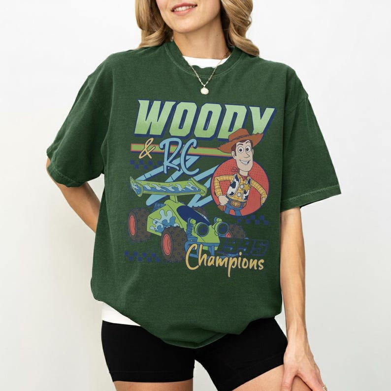 May include: Dark green t-shirt featuring a graphic of Woody and RC from Toy Story. The design includes the text "WOODY & RC Champions" in a playful font, with a cartoon image of Woody and a race car.