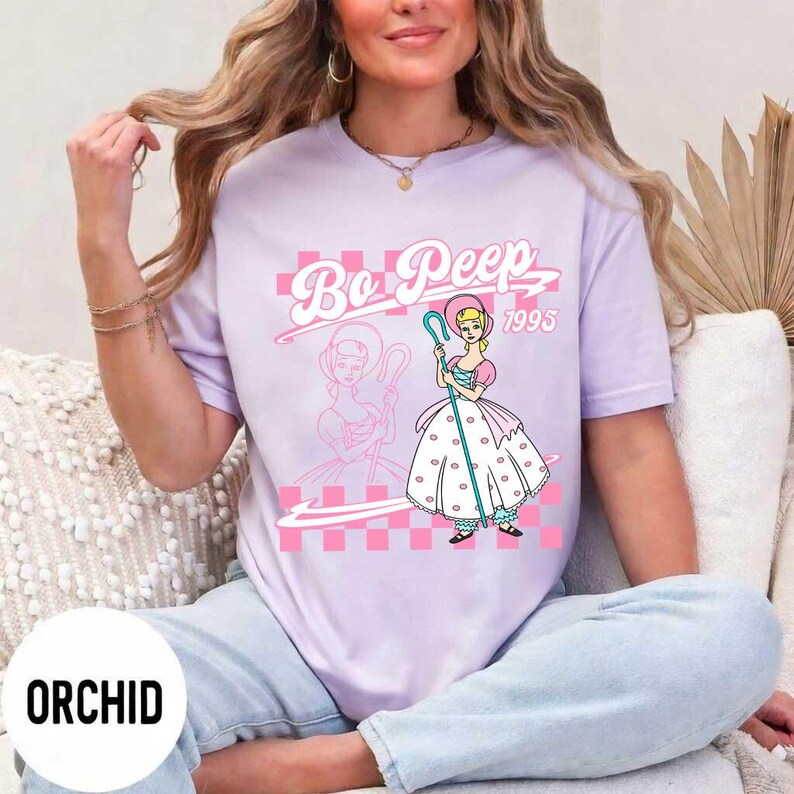 May include: A light purple t-shirt featuring a cartoon graphic of Bo Peep with the text "Bo Peep 1995" in pink. The design includes a pink checkered pattern and a silhouette of Bo Peep. The word "ORCHID" is printed at the bottom.