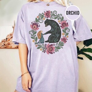 May include: A light purple t-shirt with a floral wreath design. The wreath surrounds a black bear and a person holding hands. The word "ORCHID" is in a white circle on the shirt.