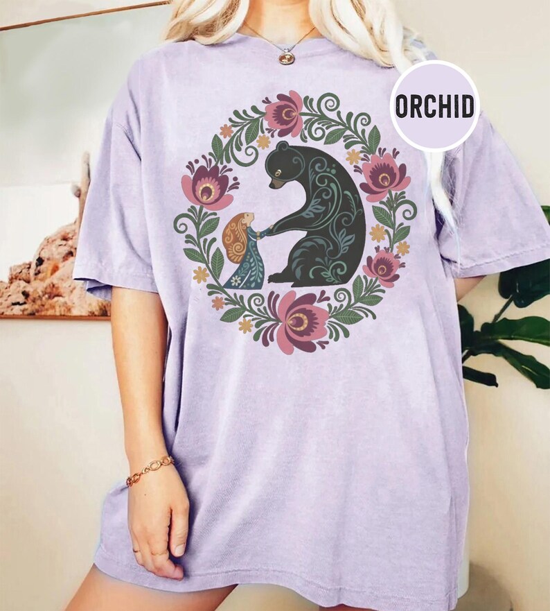 May include: A light purple t-shirt with a floral wreath design. The wreath surrounds a black bear and a person holding hands. The word "ORCHID" is in a white circle on the shirt.