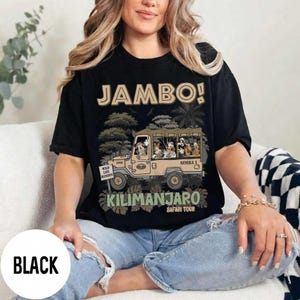 May include: Black t-shirt with a safari-themed graphic. The design features a tan jeep with cartoon characters and the words "JAMBO!" and "KILIMANJARO SAFARI TOUR". The shirt also has the text "WILD LIFE RESERVE".