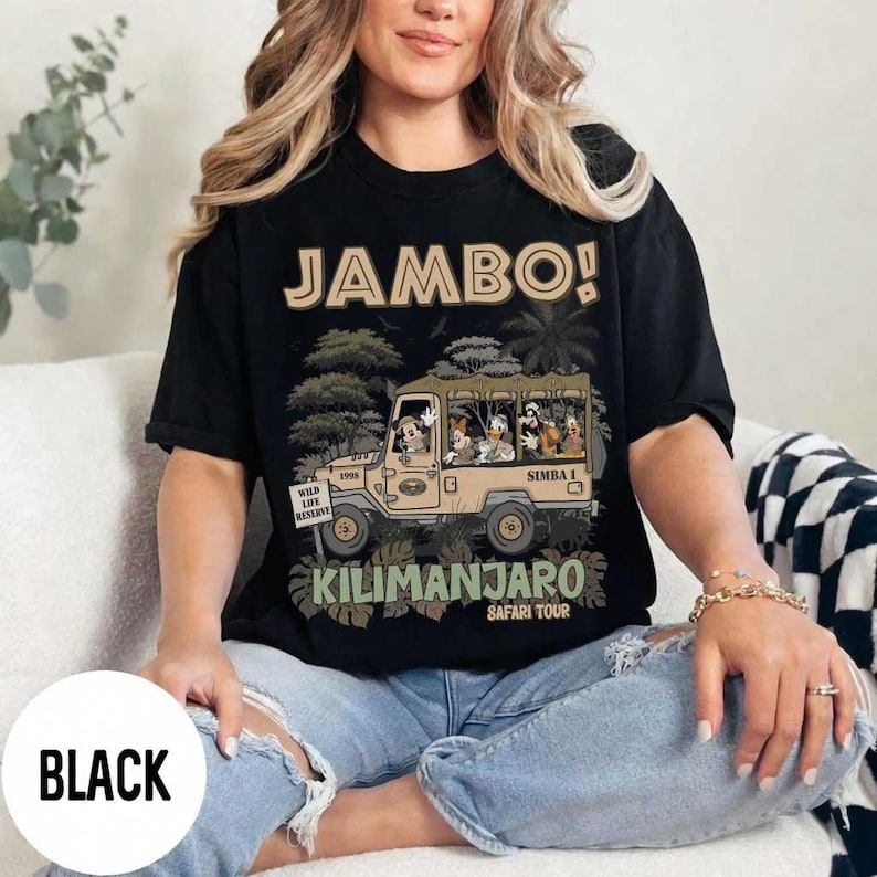 May include: Black t-shirt with a safari-themed graphic. The design features a tan jeep with cartoon characters and the words "JAMBO!" and "KILIMANJARO SAFARI TOUR". The shirt also has the text "WILD LIFE RESERVE".