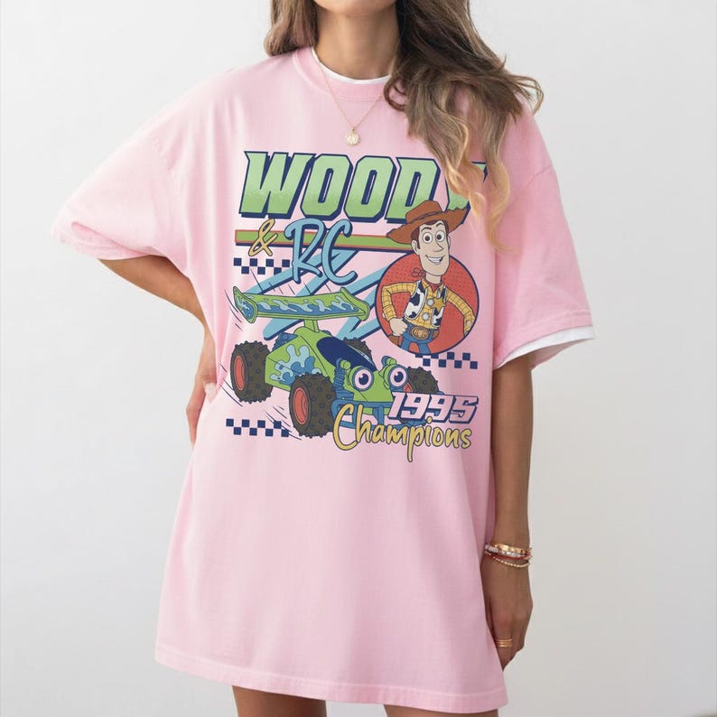May include: Pink t-shirt featuring a graphic of Woody and RC from Toy Story. The design includes the text "Woody & RC" in green and blue, a cartoon image of Woody, and the words "1995 Champions". The shirt has a white collar and sleeves.