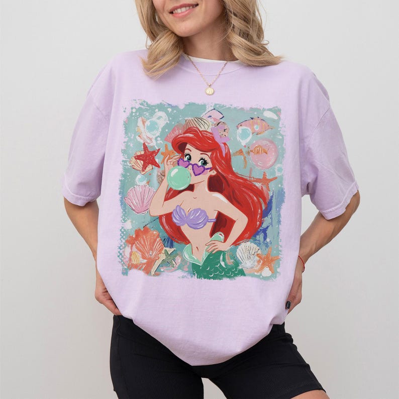 May include: A light purple short-sleeve t-shirt featuring a graphic print of a red-haired mermaid wearing heart-shaped sunglasses and blowing a green bubble. The design includes various seashells and starfish against a teal background.