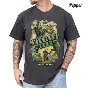 May include: A charcoal gray short-sleeve t-shirt featuring a graphic print of The Mandalorian and Grogu. The design includes the text "The Mandalorian and Grogu" and "This is the way" with illustrations of characters and vehicles in shades of green and gold.
