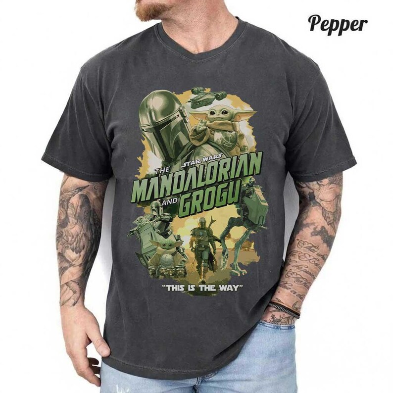 May include: A charcoal gray short-sleeve t-shirt featuring a graphic print of The Mandalorian and Grogu. The design includes the text "The Mandalorian and Grogu" and "This is the way" with illustrations of characters and vehicles in shades of green and gold.