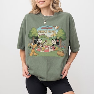 May include: Olive green t-shirt featuring a graphic of Mickey Mouse, Minnie Mouse, and Pluto having a picnic. The graphic includes the text "Mickey Mouse Perfect Picnic" in a vintage cartoon style. The shirt has short sleeves.