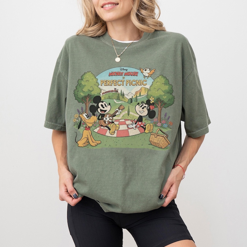 May include: Olive green t-shirt featuring a graphic of Mickey Mouse, Minnie Mouse, and Pluto having a picnic. The graphic includes the text "Mickey Mouse Perfect Picnic" in a vintage cartoon style. The shirt has short sleeves.