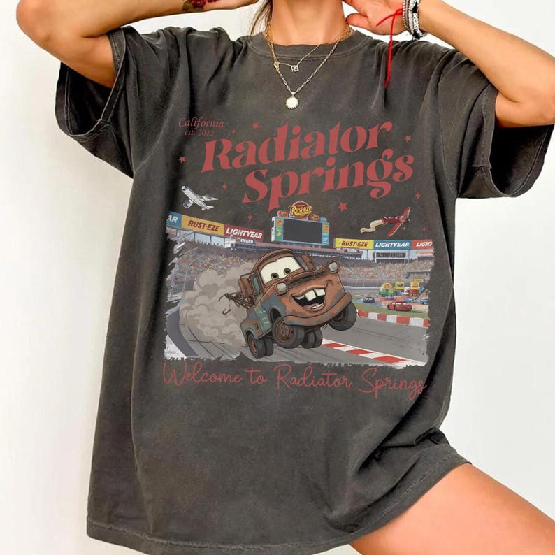 May include: Dark gray t-shirt featuring a graphic of Mater from the movie Cars, racing on a track. The shirt has the words "Radiator Springs" in red, with the text "Welcome to Radiator Springs" below the graphic. The shirt also has the text "California Est. 2012".
