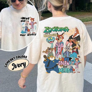 May include: Ivory-colored long-sleeve shirt featuring a graphic of characters from the animated film Zootopia. The front of the shirt has the text "Not on our watch". The back of the shirt has the text "Zootopia" and "Let's Boogie".