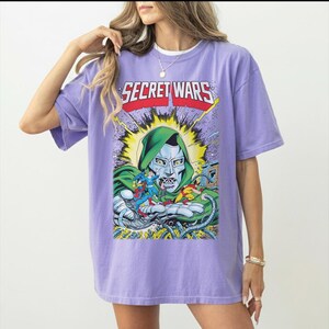 May include: A lilac-colored t-shirt featuring a comic book graphic with the text "SECRET WARS." The graphic depicts a character in a green hood and metallic mask, surrounded by other comic book characters. The shirt has short sleeves and a crew neck.