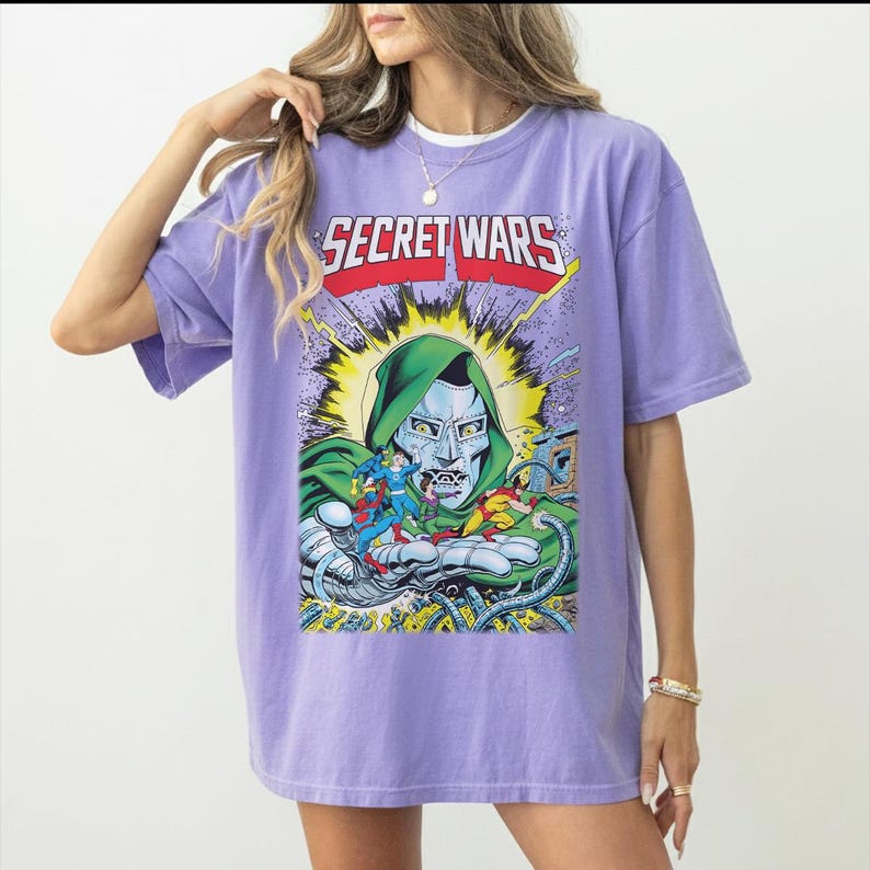 May include: A lilac-colored t-shirt featuring a comic book graphic with the text "SECRET WARS." The graphic depicts a character in a green hood and metallic mask, surrounded by other comic book characters. The shirt has short sleeves and a crew neck.