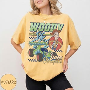 May include: Mustard-colored t-shirt featuring a graphic of Woody and RC from Toy Story. The design includes the text "WOODY & RC 1995 Champions" with a cartoon image of Woody and a green and blue RC car.