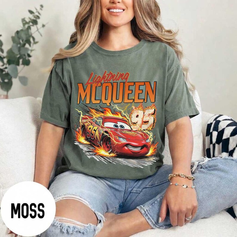 Puede incluir: Camiseta verde musgo con un dise&ntilde;o de Rayo McQueen de la pel&iacute;cula Cars. El dise&ntilde;o incluye el texto "Lightning McQueen" en naranja y un coche de carreras rojo con el n&uacute;mero "95" en llamas. La camiseta es de corte holgado.