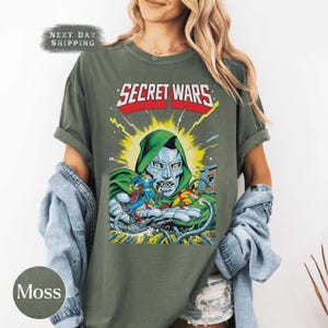 May include: Moss green t-shirt featuring a comic book graphic. The graphic includes the text "SECRET WARS" in red and white, and a depiction of Doctor Doom. The shirt has short sleeves and a relaxed fit.