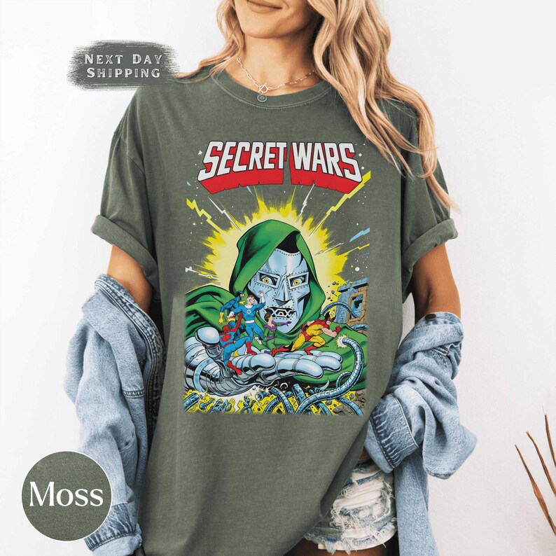 May include: Moss green t-shirt featuring a comic book graphic. The graphic includes the text "SECRET WARS" in red and white, and a depiction of Doctor Doom. The shirt has short sleeves and a relaxed fit.