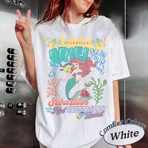 May include: A white short-sleeve t-shirt featuring a colorful graphic of Ariel the mermaid, Flounder, and Sebastian. The text reads Atlantica Ariel Ocean Tour 1991, Sebastian with the Hot Crustacean Band, with coral and seaweed illustrations.