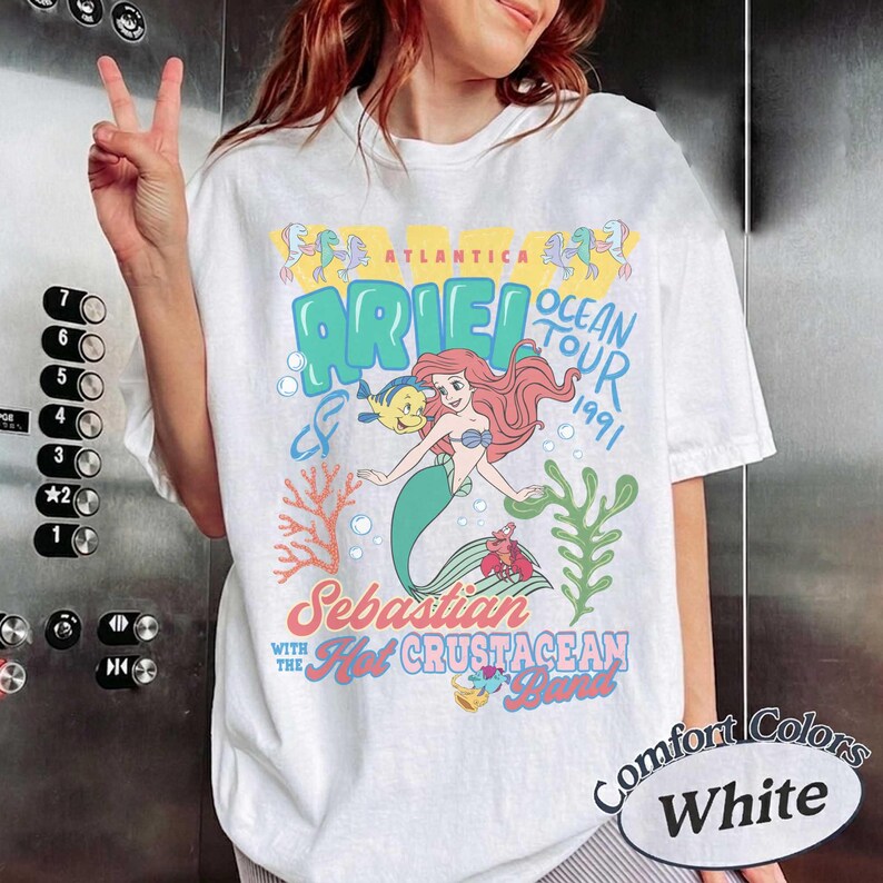 May include: A white short-sleeve t-shirt featuring a colorful graphic of Ariel the mermaid, Flounder, and Sebastian. The text reads Atlantica Ariel Ocean Tour 1991, Sebastian with the Hot Crustacean Band, with coral and seaweed illustrations.