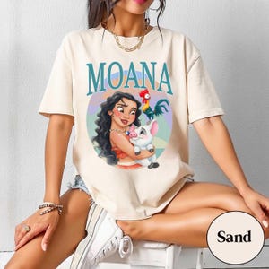 May include: Sand-colored t-shirt featuring the word "MOANA" in teal above a colorful illustration of Moana, a pig, and a rooster. The graphic is set within a circular design. The shirt is short-sleeved.