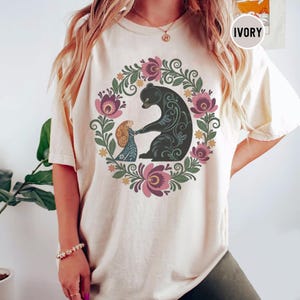 May include: Ivory-colored t-shirt featuring a floral wreath design with a black bear and a girl holding hands. The wreath includes pink and purple flowers and green leaves. The word "IVORY" is printed on the top right.