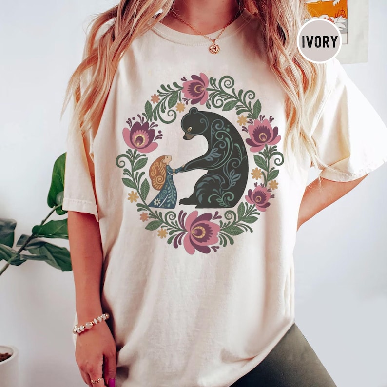 May include: Ivory-colored t-shirt featuring a floral wreath design with a black bear and a girl holding hands. The wreath includes pink and purple flowers and green leaves. The word "IVORY" is printed on the top right.