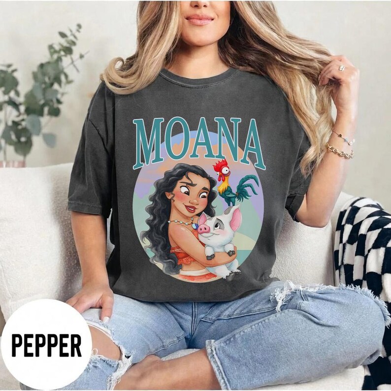 May include: Dark gray t-shirt featuring the word "MOANA" in teal lettering above a colorful graphic. The graphic depicts a cartoon woman holding a pig, with a rooster nearby. The shirt is a relaxed fit.
